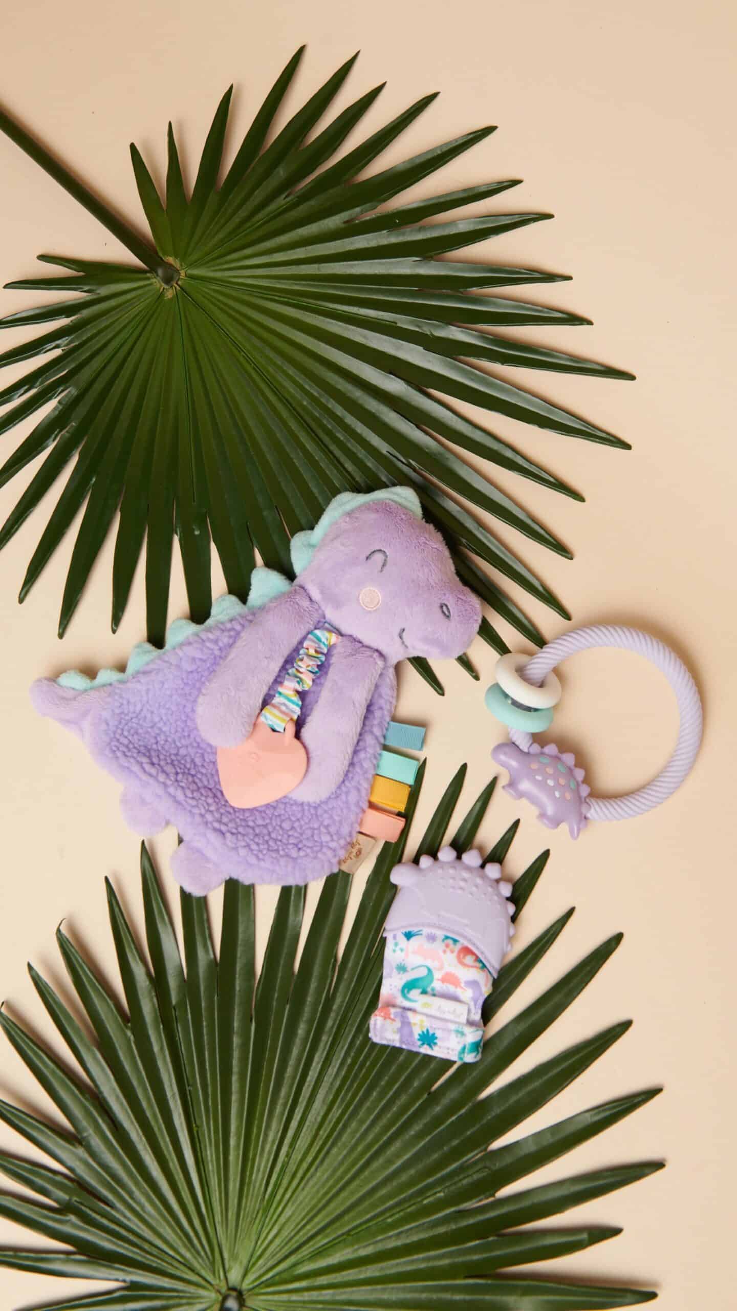 itzy-ritzy-lov8430-itzy-lovey-purple-din A plush purple dinosaur toy, a baby teething ring, and a baby mitten with rainbow designs are placed on green palm leaves set against a beige background.