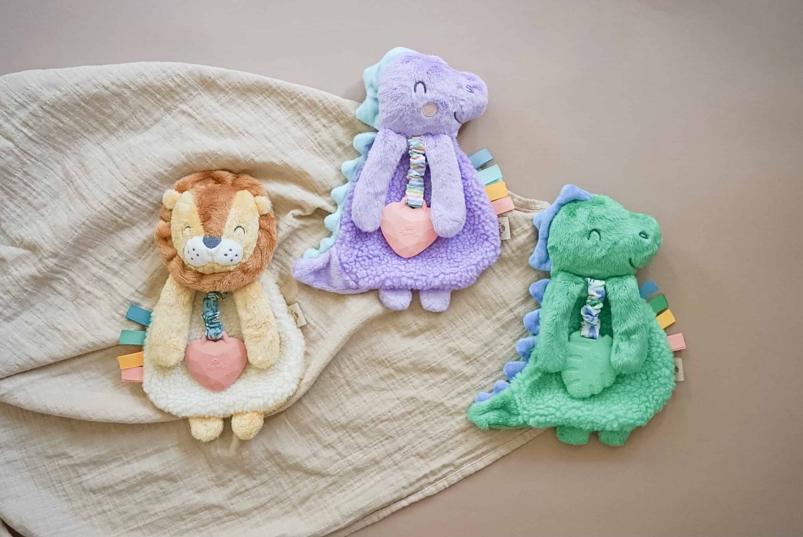 itzy-ritzy-lov8430-itzy-lovey-purple-din Three plush toys, a lion, a lavender dinosaur, and a green dinosaur, are placed on a beige blanket on a neutral background. Each toy holds a heart-shaped object and has colorful tags on its sides.