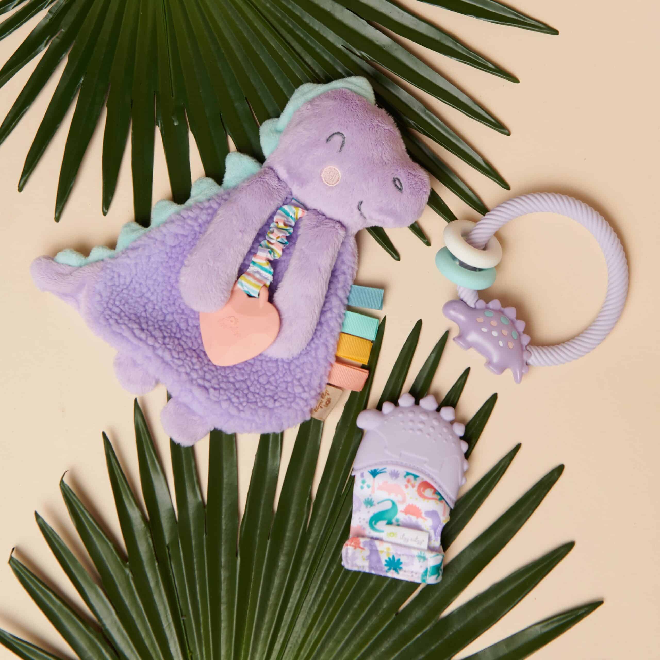 itzy-ritzy-lov8430-itzy-lovey-purple-dino-1 A purple dinosaur plush toy, a lavender teething ring with beads, and a dinosaur-themed baby mitten are arranged on palm leaves.