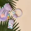 A collection of baby toys: a purple plush dinosaur, a teething ring with colorful beads, and a pacifier clip with a dinosaur design, all placed on green palm leaves against a beige background.