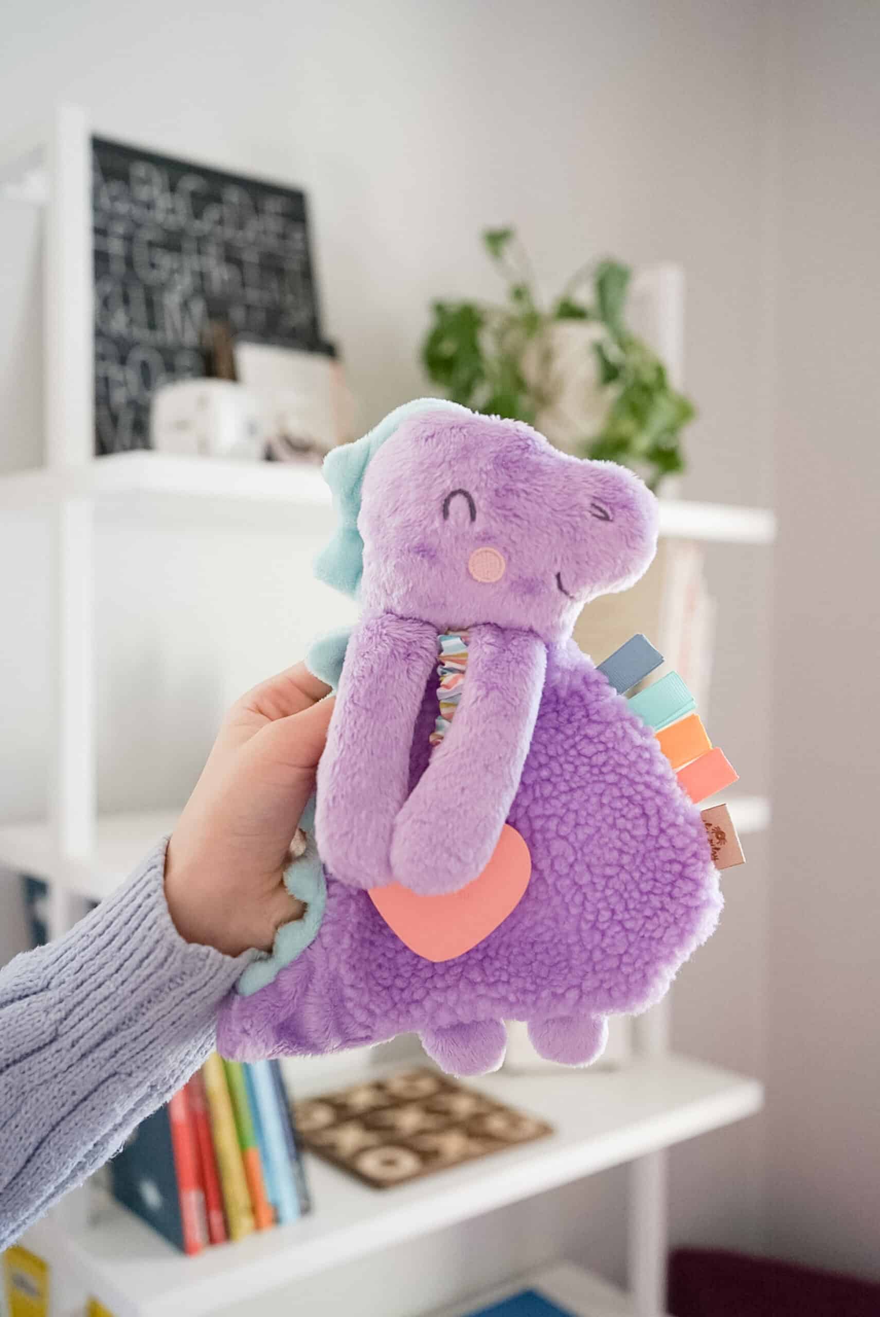 itzy-ritzy-lov8430-itzy-lovey-purple-dino-2 A hand is holding a purple stuffed toy shaped like a dinosaur with a small orange heart on its belly. The background shows white shelves with books and a plant.
