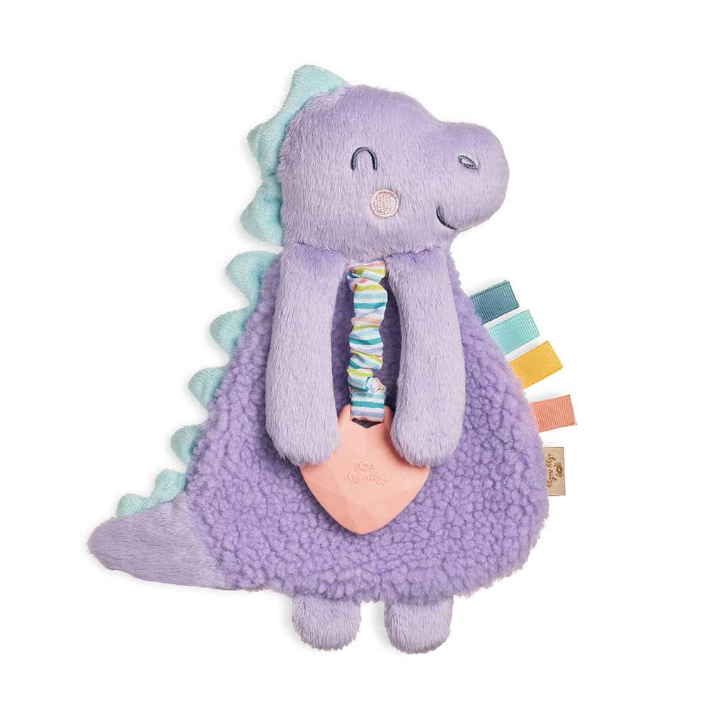 itzy-ritzy-lov8430-itzy-lovey-purple-dino-3 Plush purple dinosaur toy with a soft blue spine, holding a rubber heart-shaped teething ring. It has various colorful fabric tags attached to its body.