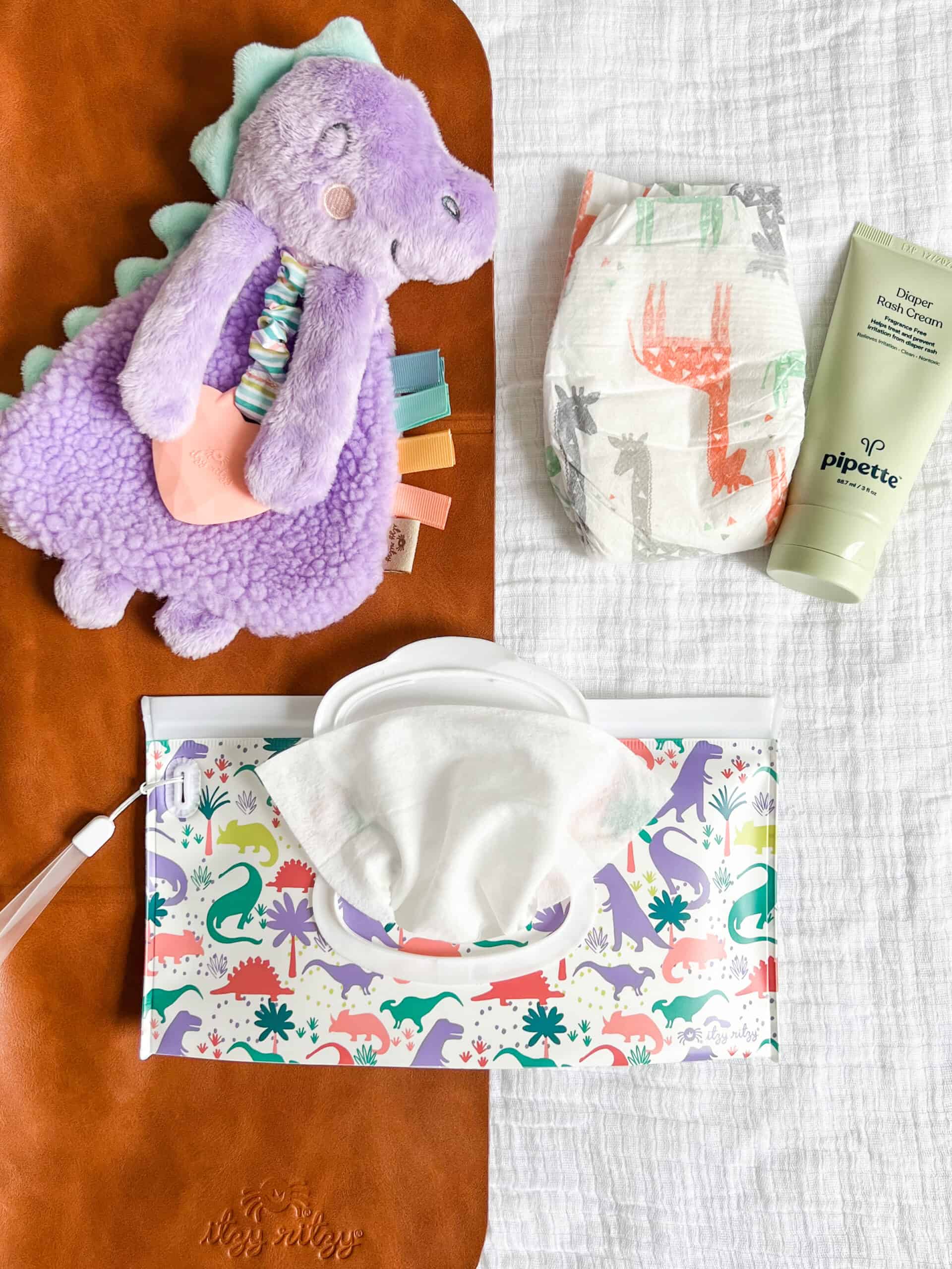 itzy-ritzy-lov8430-itzy-lovey-purple-dino-6 A flat lay of baby items: a purple dinosaur plush toy, a diaper with animal prints, a tube of pipette baby balm, a pack of wipes, and a brown changing pad all placed on a white cloth.