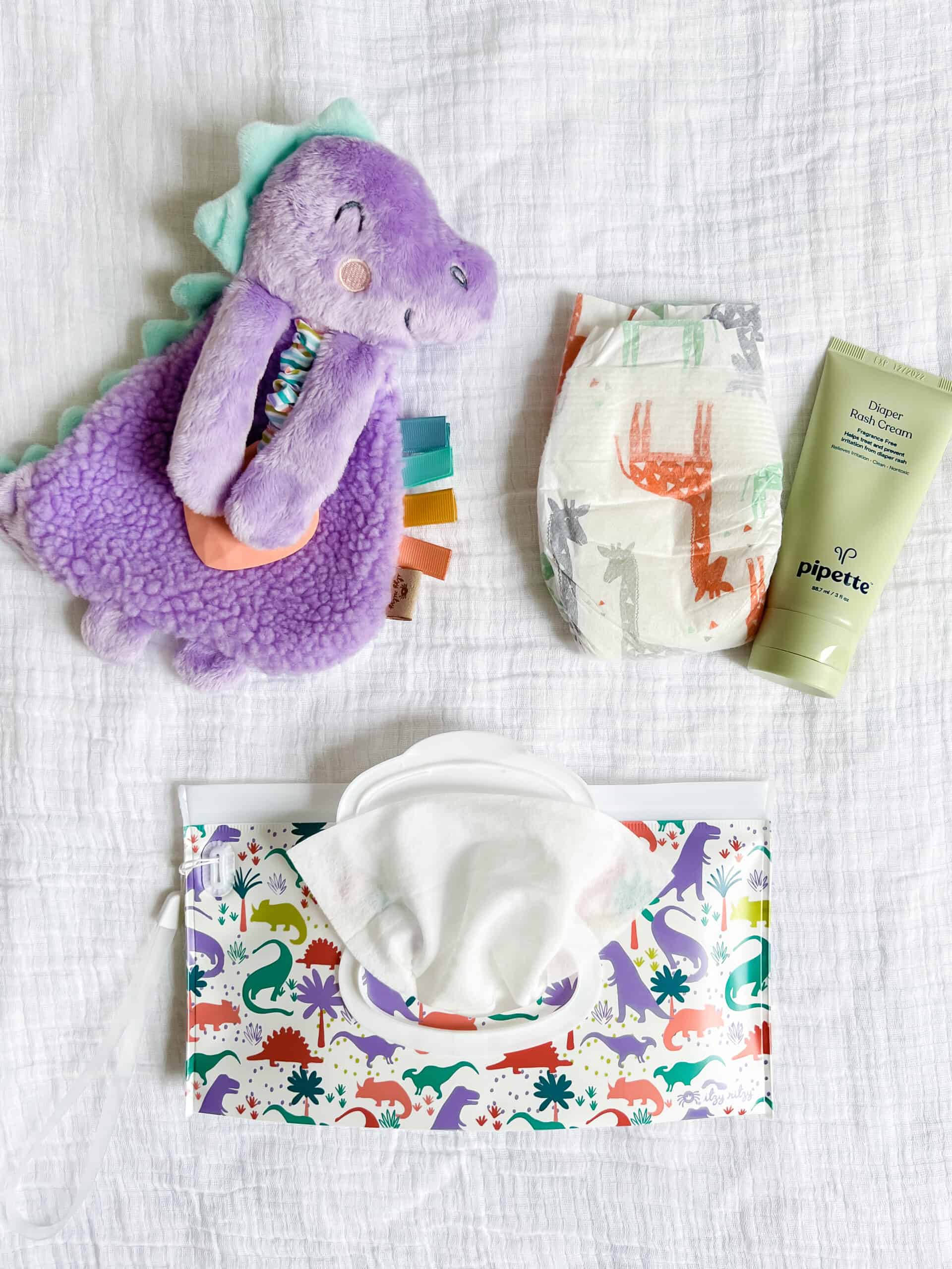 itzy-ritzy-lov8430-itzy-lovey-purple-dino-7 A purple plush dinosaur toy, a diaper with dinosaur prints, a tube of Pipette baby cream, and a pack of dinosaur-themed baby wipes on a white fabric background.
