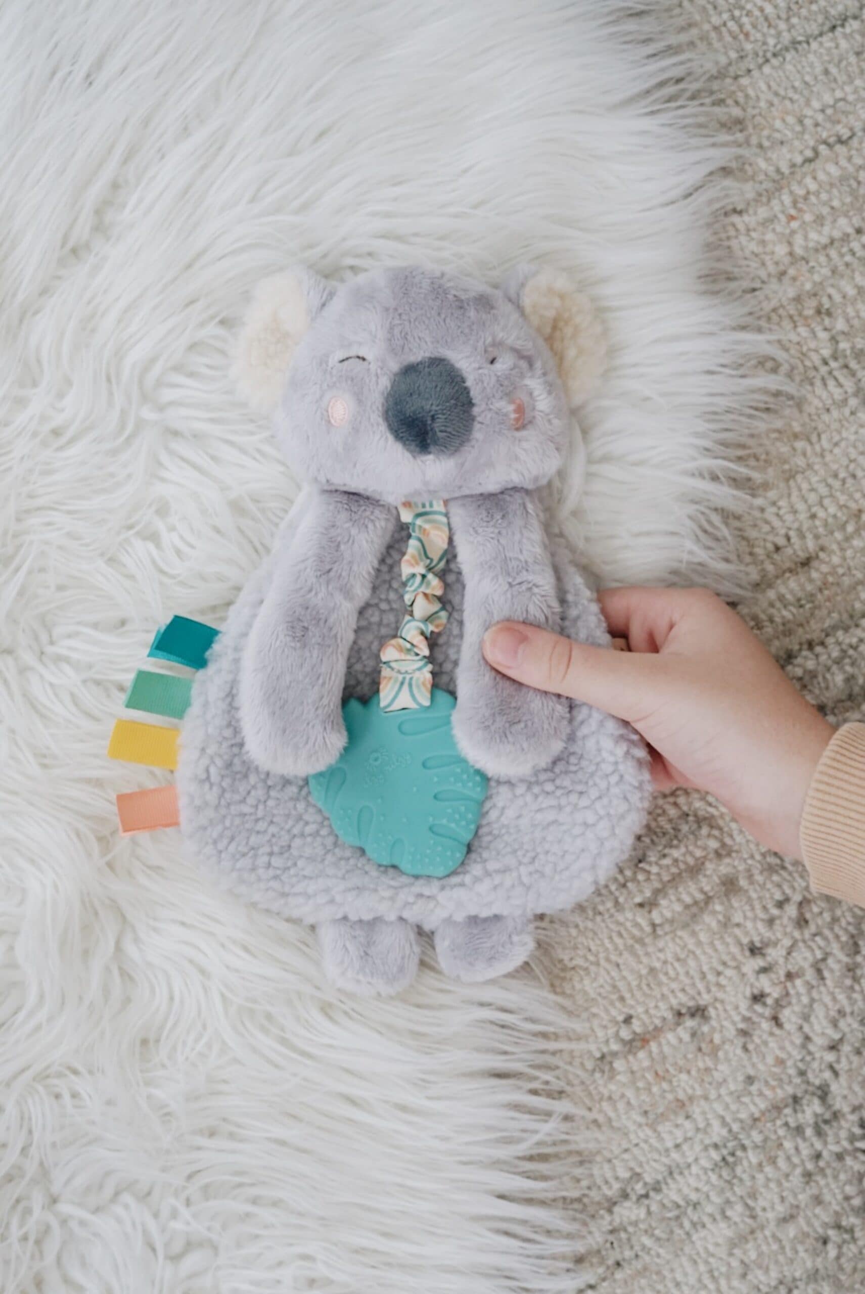 Processed with VSCO with a6 preset A hand holding a plush koala toy with a teal teether attached to its front and colorful fabric tabs on its side, lying on a fluffy white surface.