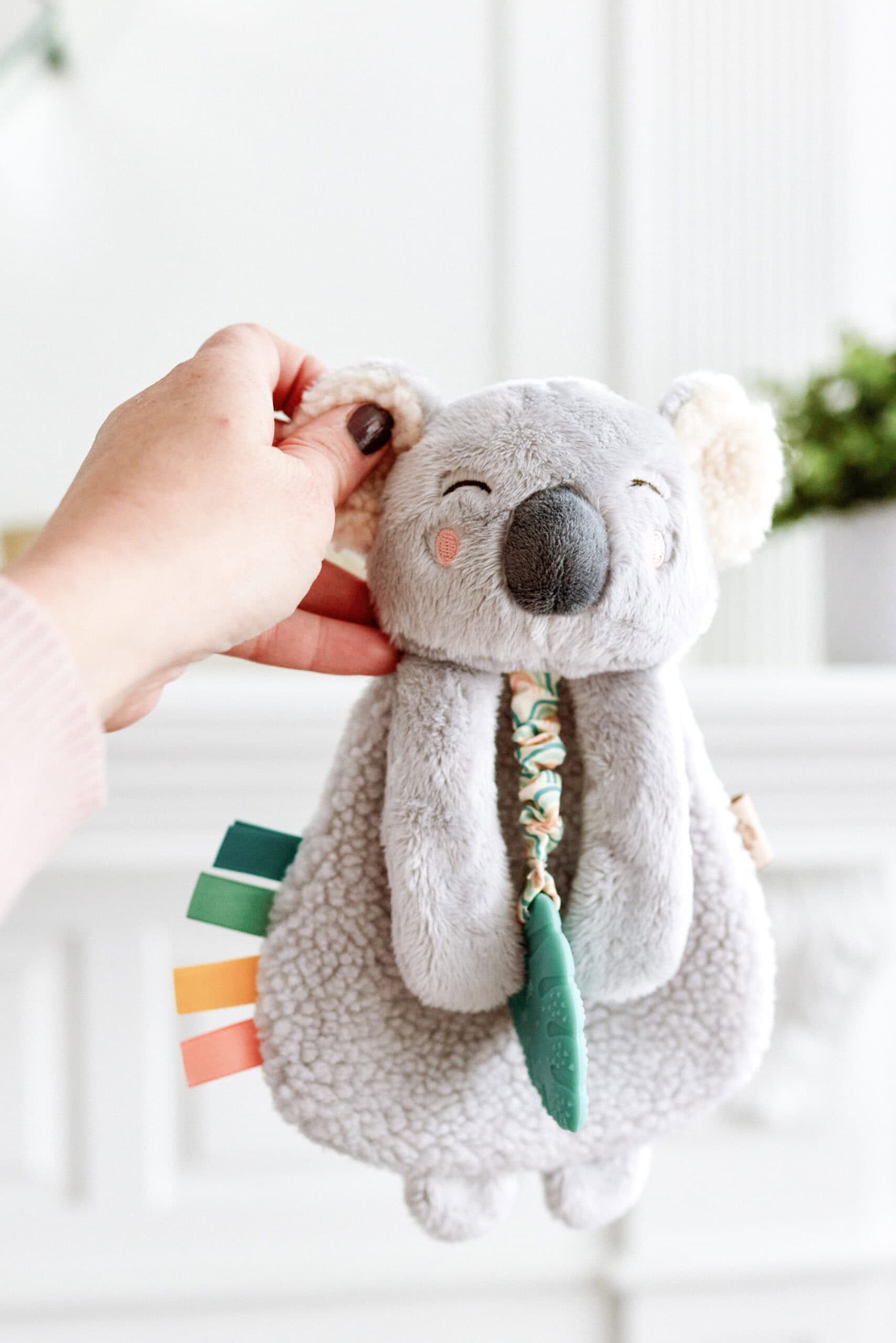 itzy-ritzy-lov8433-itzy-lovey-koala-8 A hand holding a gray, plush koala toy with colorful ribbon tags and a textured blue teething ring, against a light-colored background.
