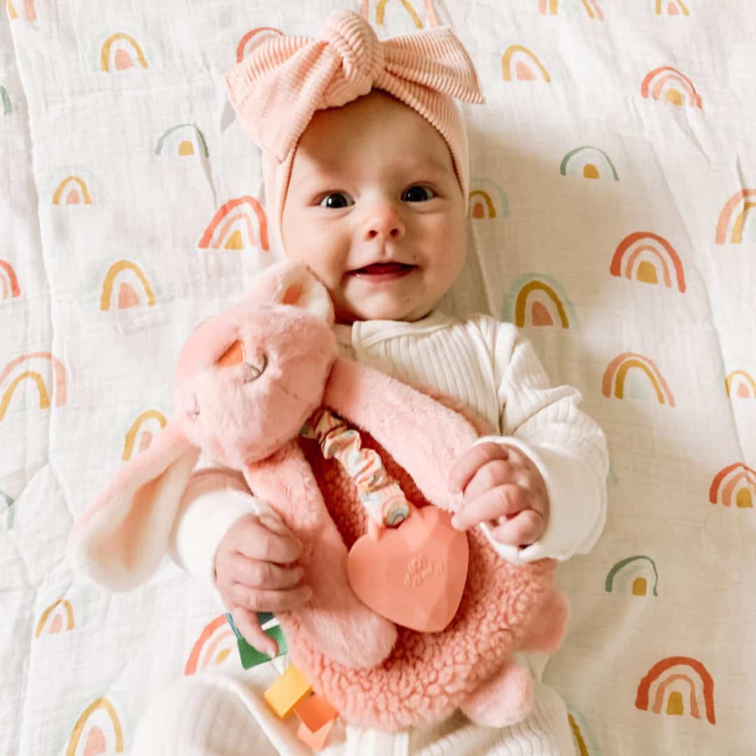 itzy-ritzy-lov8447-itzy-lovey-bunn A baby dressed in white with a pink headband smiles while holding a pink stuffed animal. The background features a white blanket with colorful rainbow patterns.