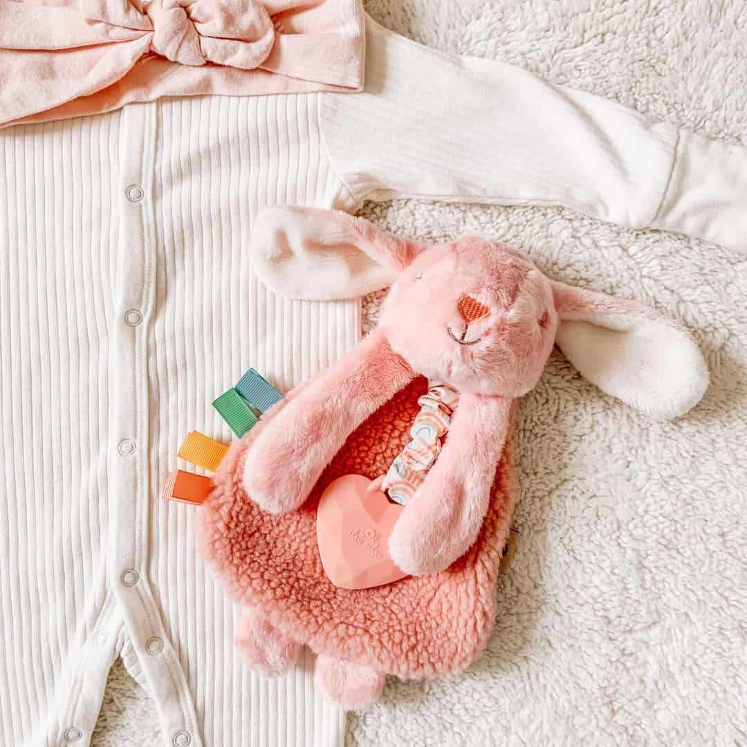 itzy-ritzy-lov8447-itzy-lovey-bunny-1 A pink plush rabbit toy and a white ribbed baby onesie with buttons laying on a soft textured surface. A pink headband is partially visible at the top of the image.