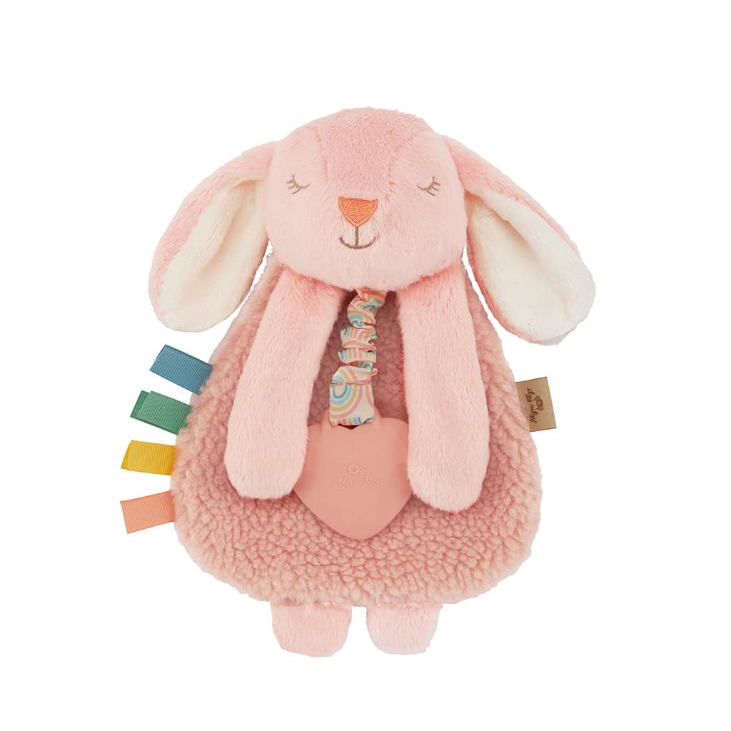 itzy-ritzy-lov8447-itzy-lovey-bunny-1 A soft, pink plush bunny toy with long ears and various colorful fabric tags attached to its side, holding a heart-shaped accessory.