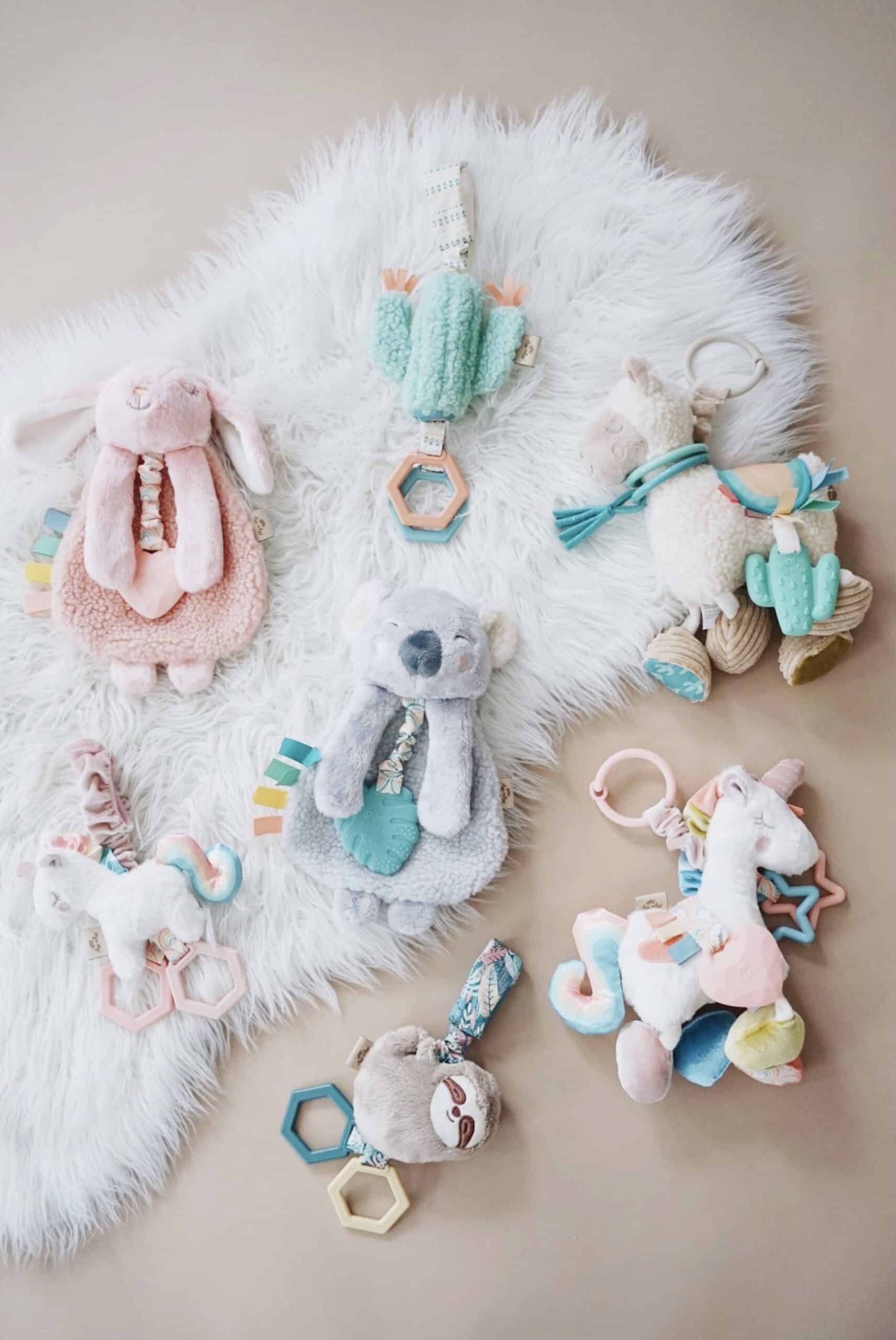Processed with VSCO with a6 preset A collection of soft plush baby toys on a white fuzzy surface, including pastel-colored animals such as a rabbit, koala, sloth, and unicorn, each with attached teething rings and colorful fabrics.