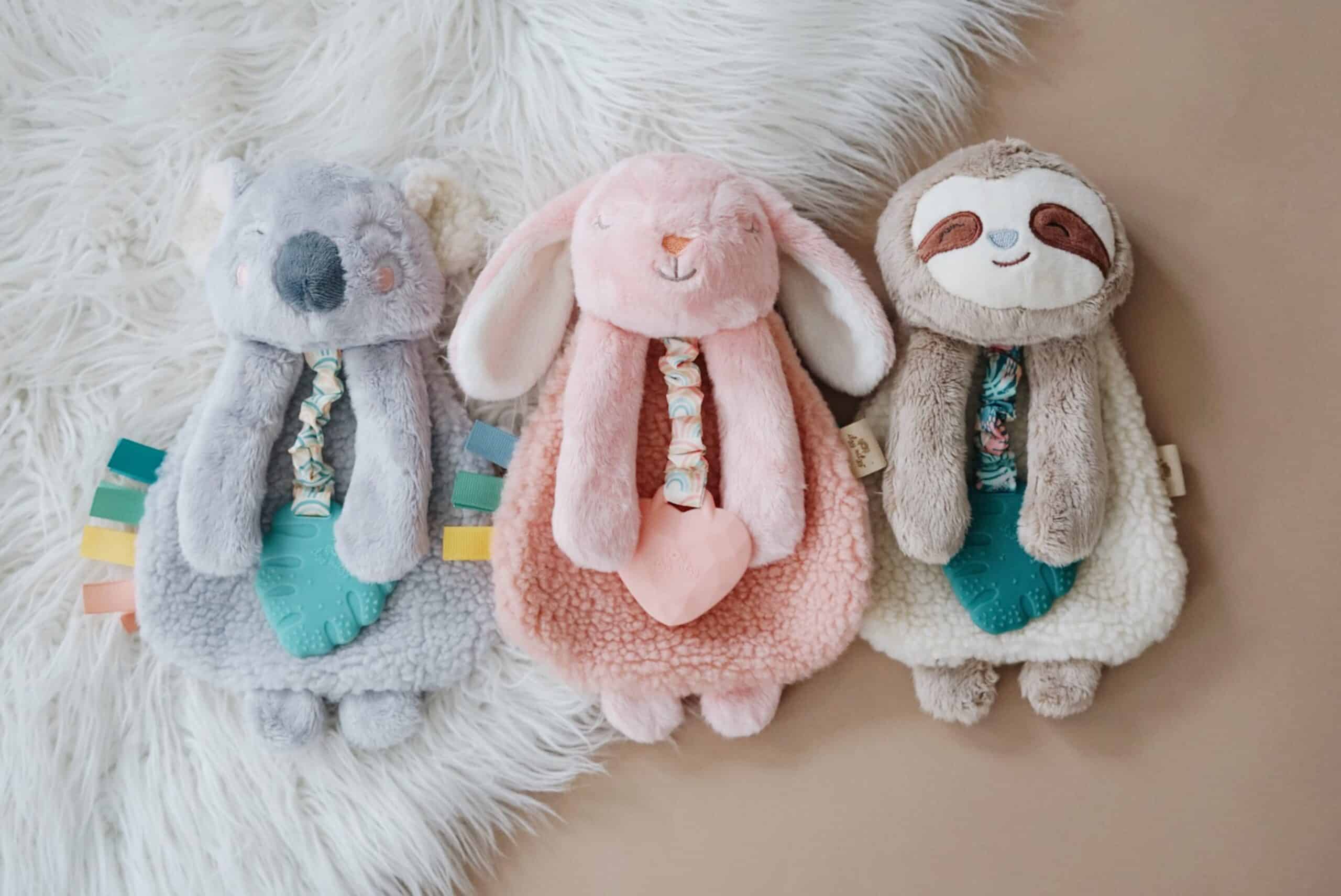 Processed with VSCO with a6 preset Three plush toys sit side by side on a fluffy white rug. From left to right, they resemble a koala, a pink rabbit, and a sloth. Each toy holds a soft fabric heart and colorful tags.