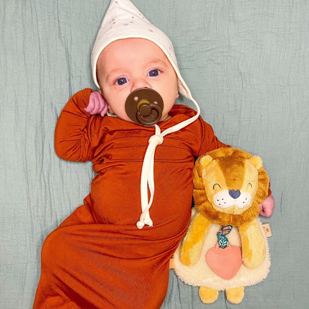 itzy-ritzy-lov8472-itzy-lovey-lio An infant in an orange sleeper and white hat lies on a green blanket, holding a stuffed lion toy. The baby has a pacifier in their mouth.