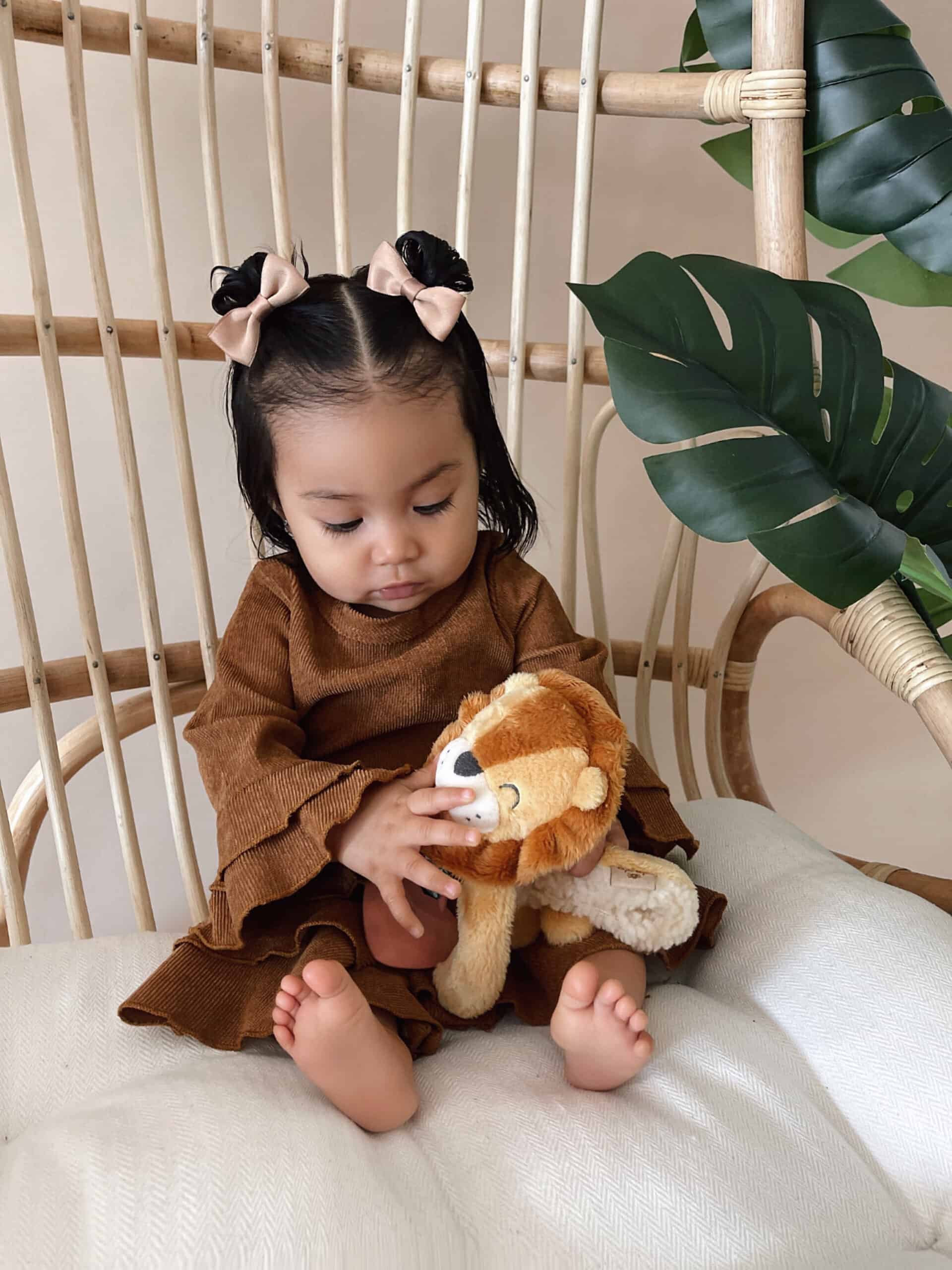 itzy-ritzy-lov8472-itzy-lovey-lion-10 A toddler girl sits on a wicker chair holding a stuffed lion toy, dressed in a brown outfit with hair styled in two buns with bows. Large green leaves decorate the background.
