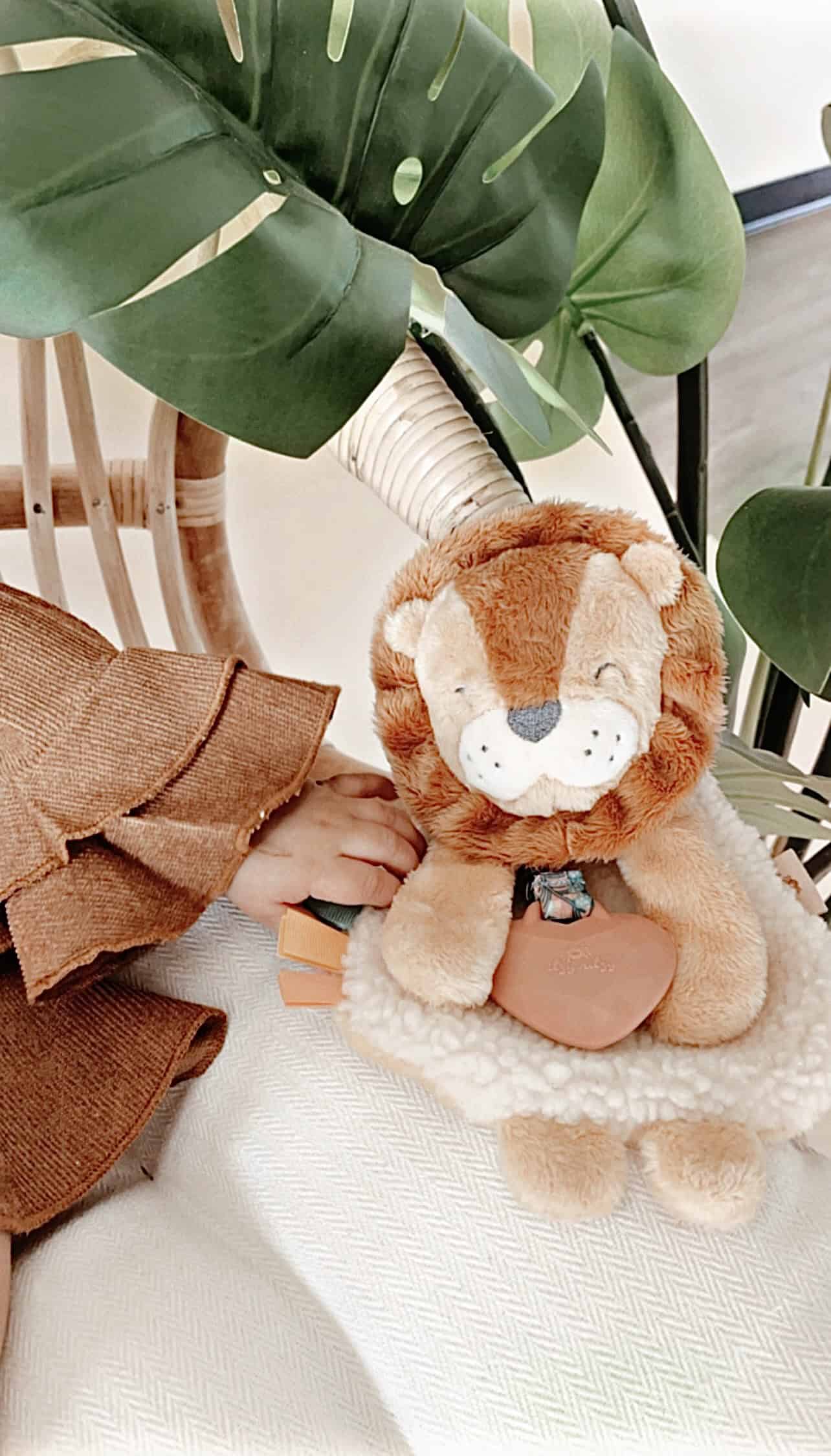 itzy-ritzy-lov8472-itzy-lovey-lion-12 A plush lion toy is placed on a white surface next to a child’s hand and large green leaves. The lion is holding a small object in its front paws.