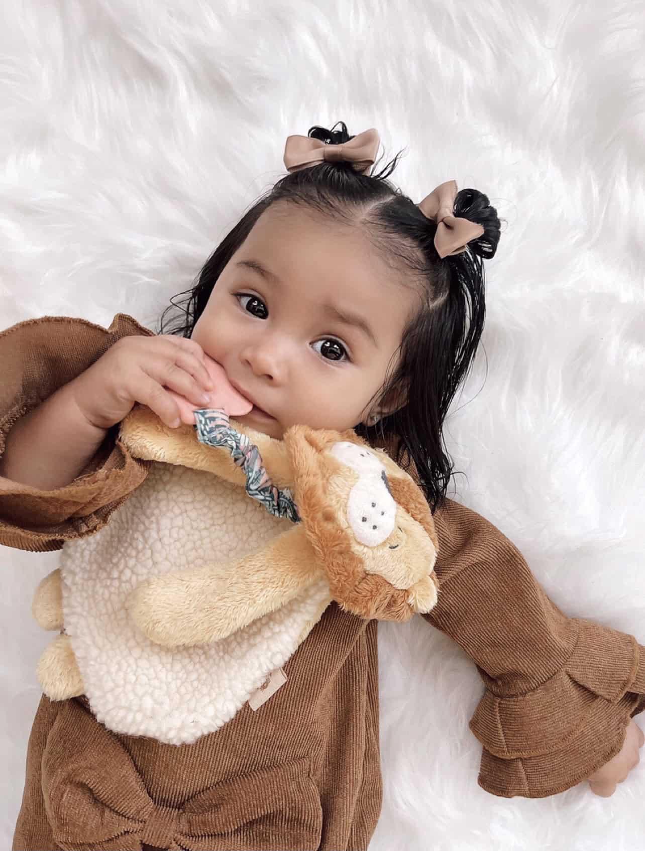 itzy-ritzy-lov8472-itzy-lovey-lion-13 A baby with brown hair and bows lies on a white fluffy surface, wearing a brown outfit and holding a plush lion teether.