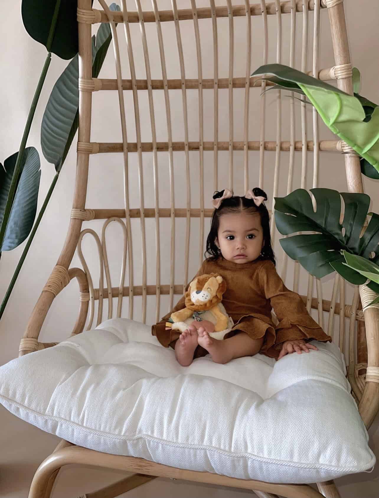 itzy-ritzy-lov8472-itzy-lovey-lion-14 A baby sits on a cushioned wicker chair holding a stuffed lion toy, surrounded by large green leaves.