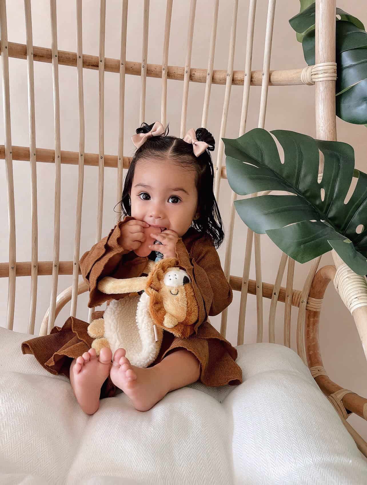 itzy-ritzy-lov8472-itzy-lovey-lion-15 A toddler with two small bows in her hair sits on a wicker chair holding a stuffed animal, surrounded by green plant leaves.