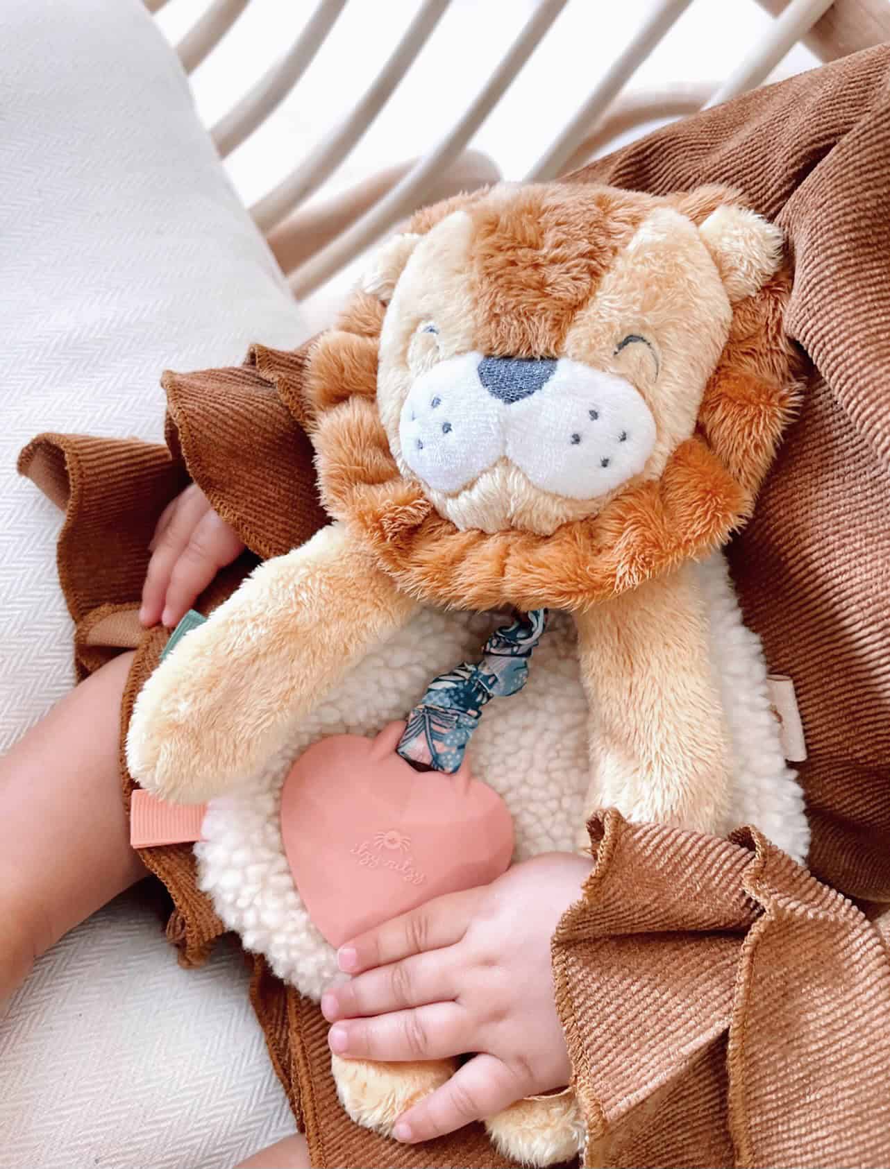 itzy-ritzy-lov8472-itzy-lovey-lion-16 A child holds a plush lion toy with a pink heart-shaped teether and wears a brown outfit with ruffled sleeves.