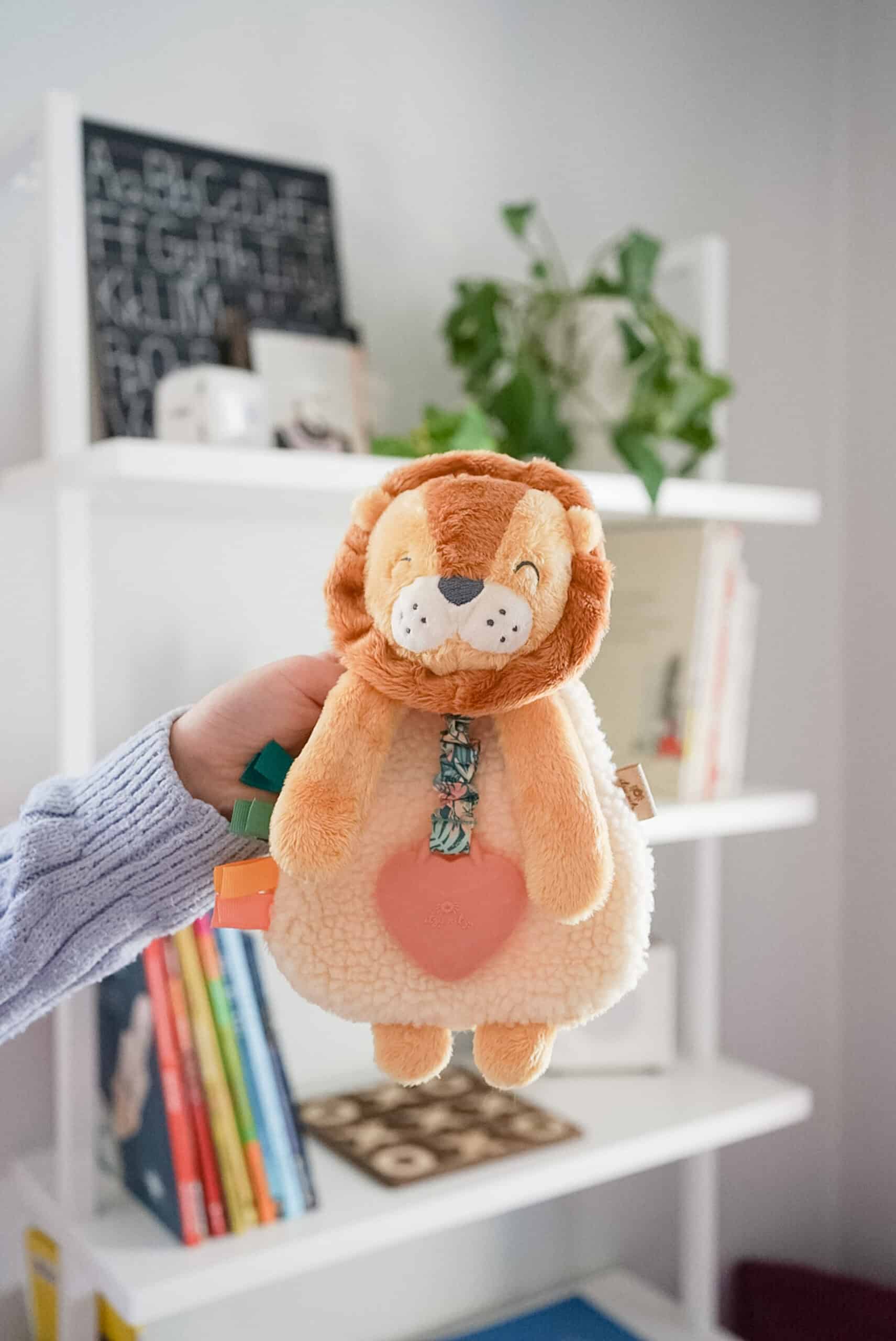 itzy-ritzy-lov8472-itzy-lovey-lion-5 A person holding a plush lion toy in front of a white bookshelf containing books, a potted plant, and decorative items.