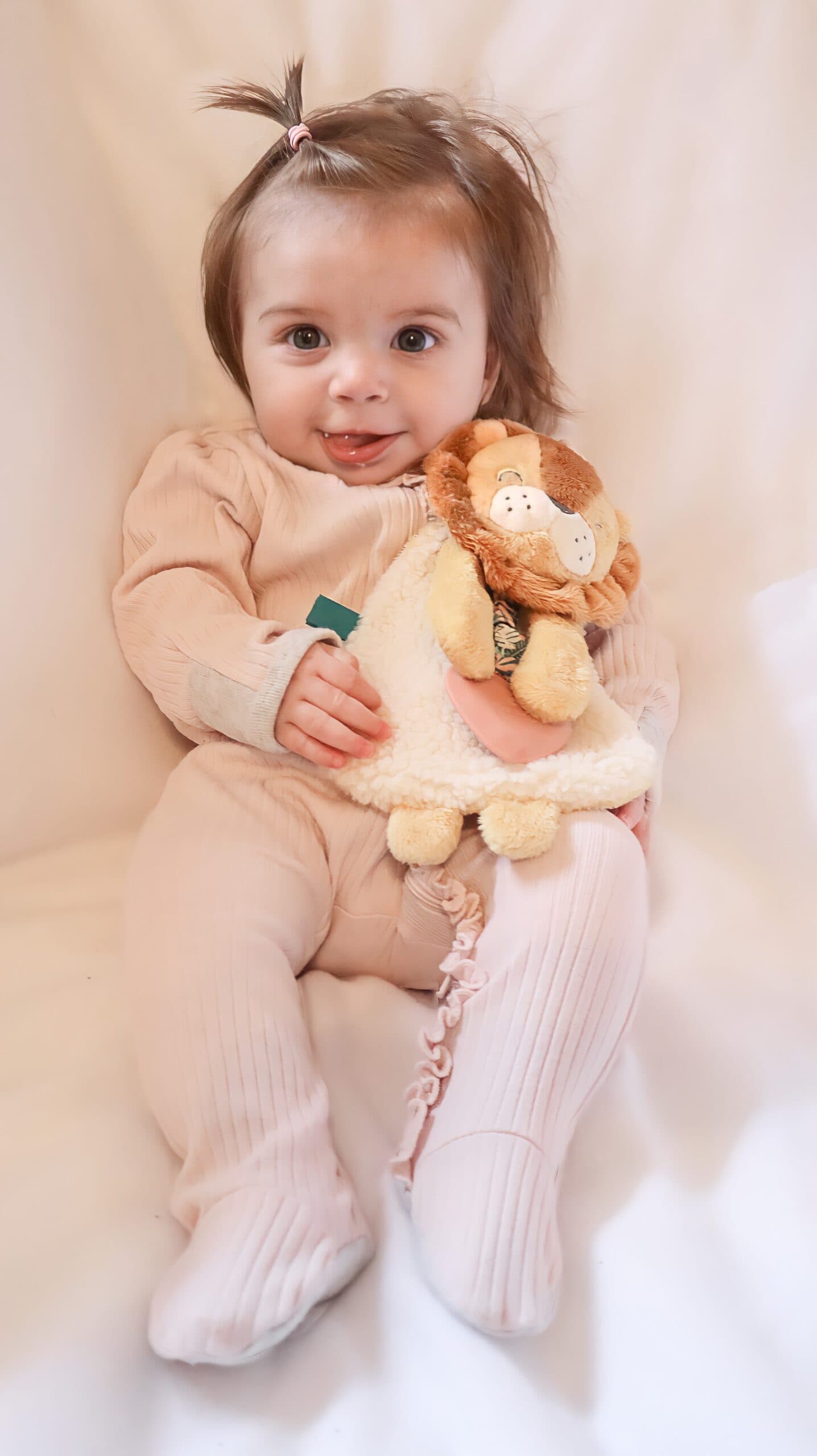 itzy-ritzy-lov8472-itzy-lovey-lion-7 A baby in light pink clothing sits on a white surface, holding a stuffed lion toy.