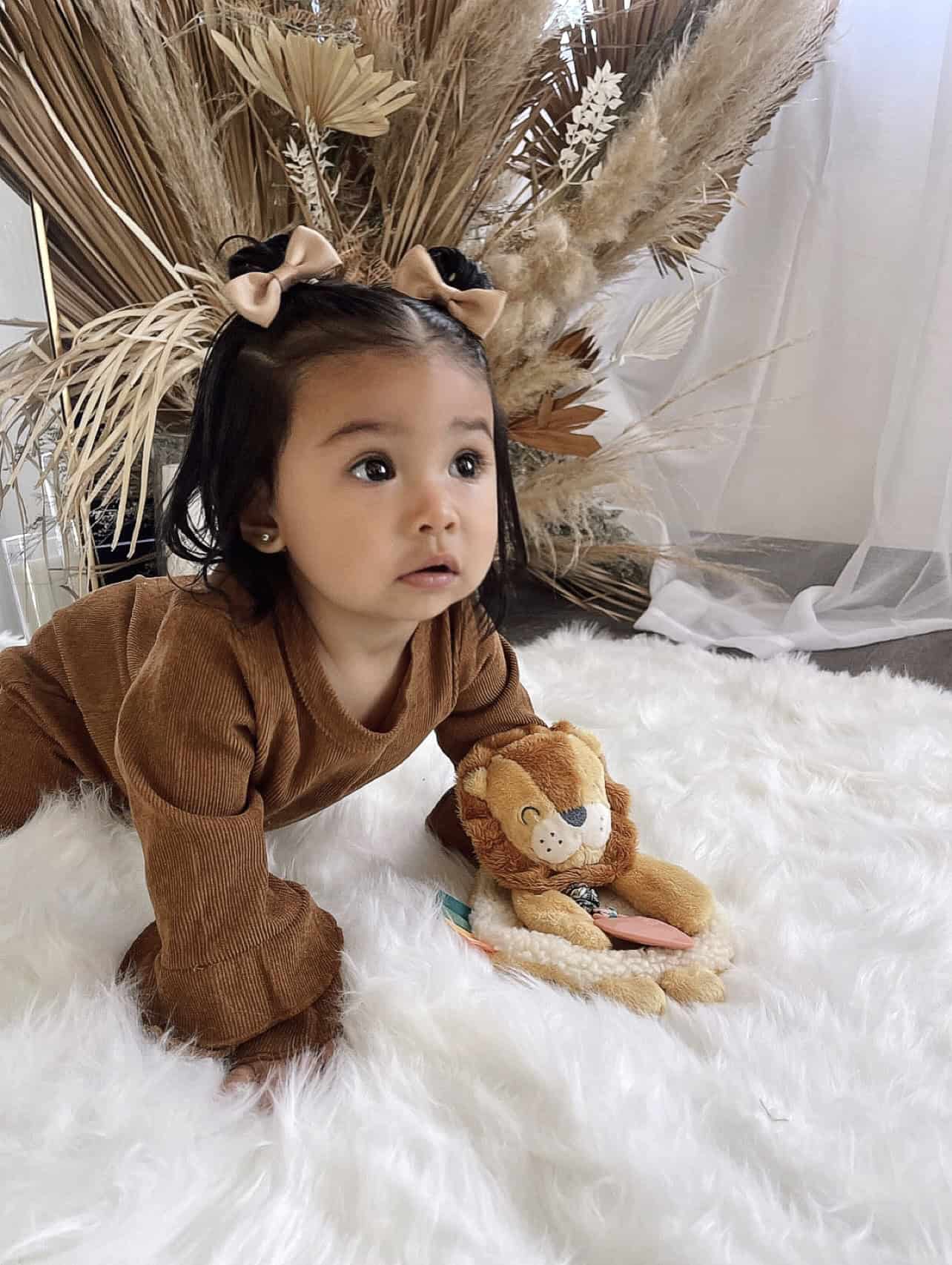 itzy-ritzy-lov8472-itzy-lovey-lion-9 A toddler with bows in her hair and wearing a brown outfit is crawling on a white fluffy rug. She is holding a plush lion toy. Dried brown plants and sheer curtains are in the background.