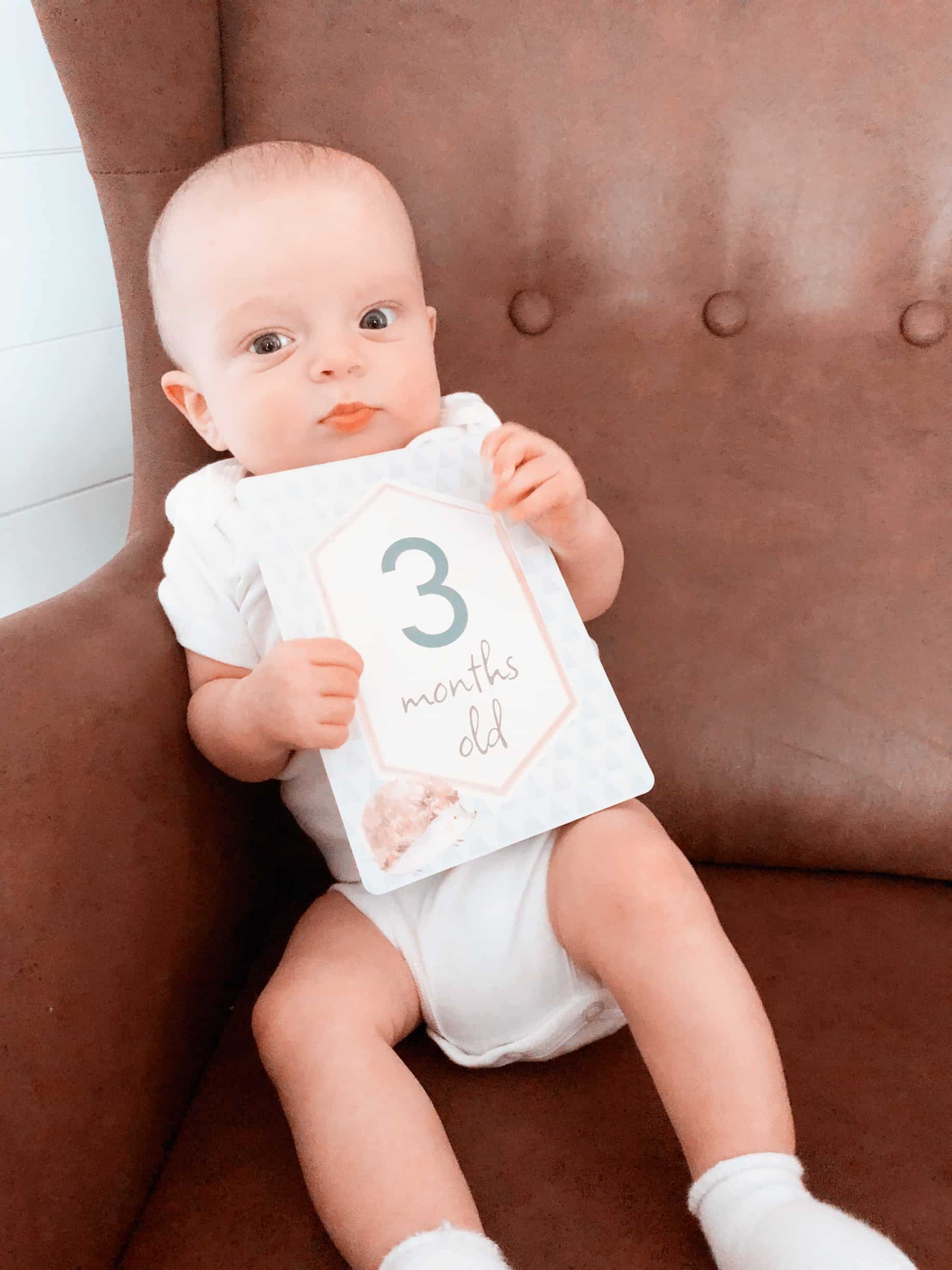 itzy-ritzy-mc8383-itzy-moments-woodland-2 A baby is sitting in a brown chair holding a number sign.