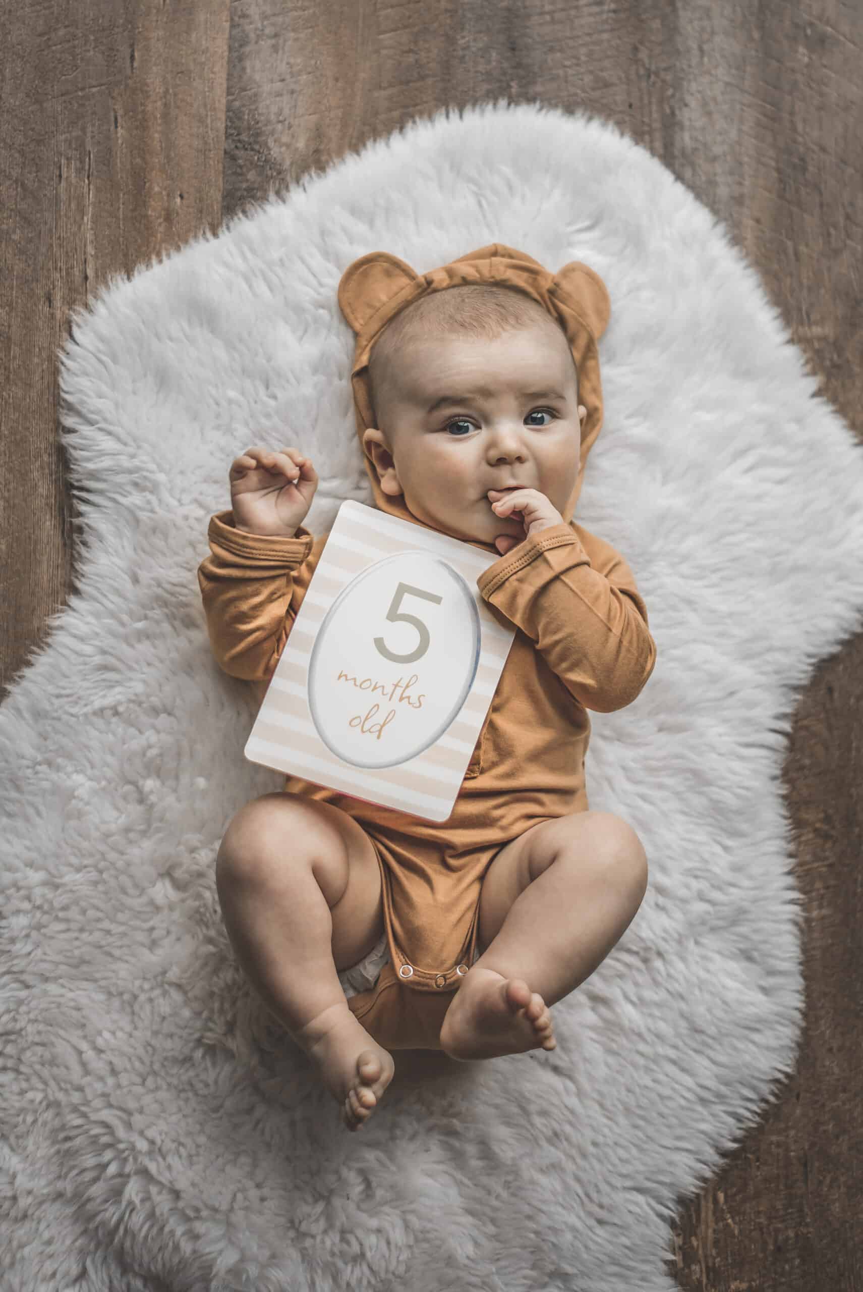 itzy-ritzy-mc8383-itzy-moments-woodland-3 A baby wearing a teddy bear holding a number sign.