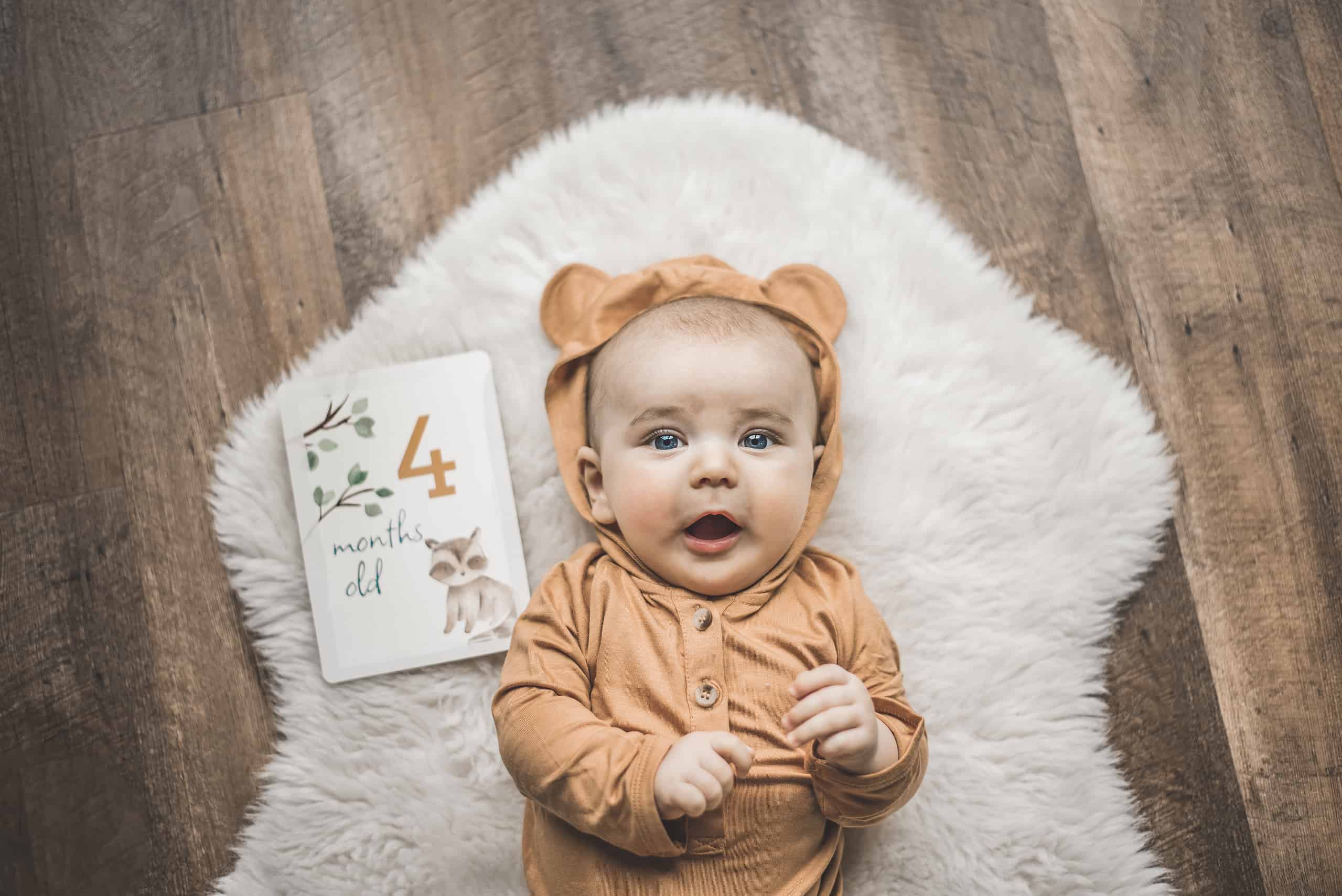 itzy-ritzy-mc8383-itzy-moments-woodland-5 A baby laying on a rug with a book and a teddy bear.