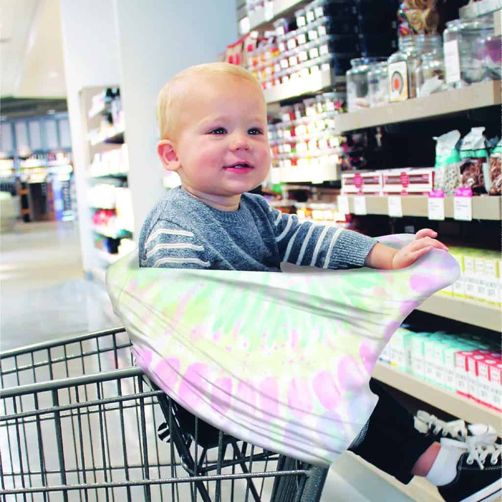 A baby sitting in a Mom Boss™ 4-IN-1 Multi-Use Nursing Cover & Scarf in a grocery store.