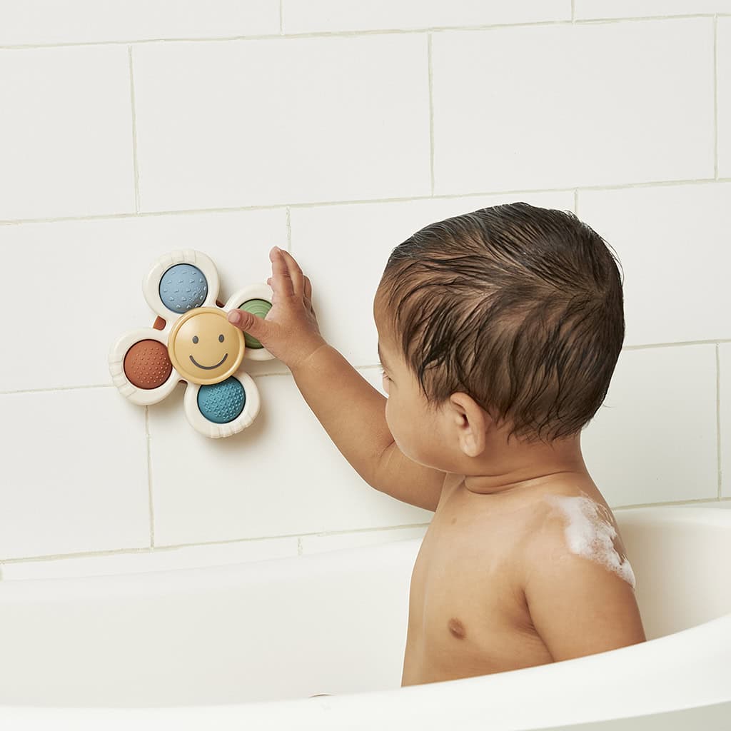 itzy-ritzy-spin8547-itzy-pop-&-whirl-smi A baby playing with a flower-shaped bath toy attached to a tiled bathroom wall.