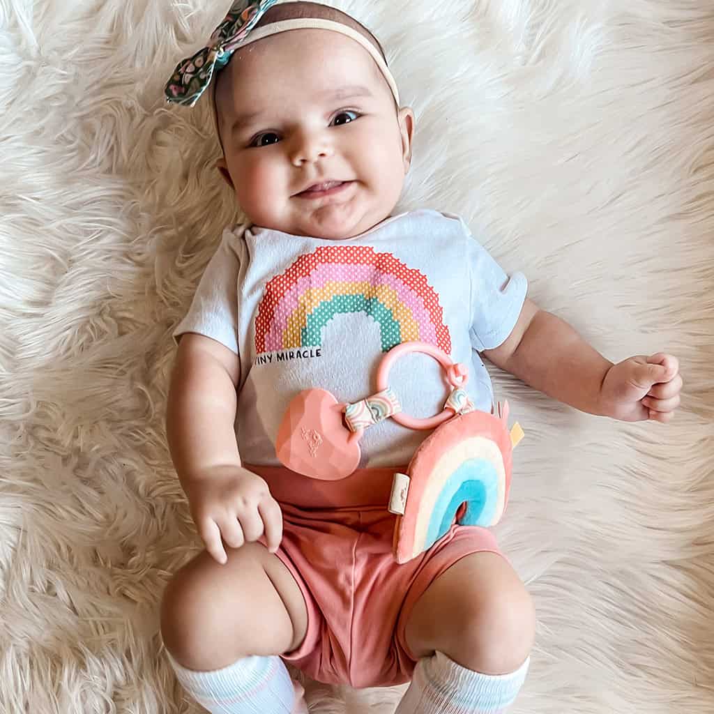 itzy-ritzy-tpl3003-itzy-pal-rainbow-1 A smiling baby lying on a fluffy rug, dressed in a light gray shirt and peach shorts, holding an Itzy Ritzy - Itzy Pal - Plush Pal Infant Toy with Silicone Teether, with a bib that reads "tiny miracle.