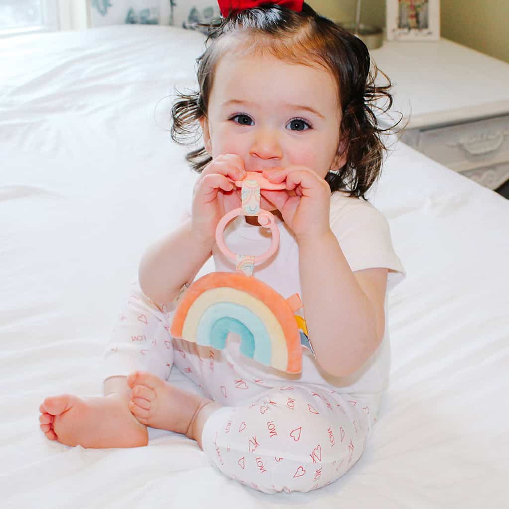 itzy-ritzy-tpl3003-itzy-pal-rainbow-2 A toddler with curly hair, wearing a rainbow-print shirt and patterned pants, sits on a bed and chews on an Itzy Ritzy - Itzy Pal - Plush Pal Infant Toy with Silicone Teether.