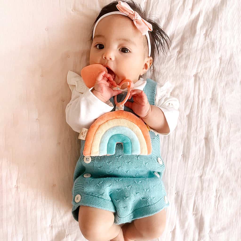 itzy-ritzy-tpl3003-itzy-pal-rainbow-7 Baby with a headband lying on a pink blanket, chewing on an Itzy Ritzy - Itzy Pal - Plush Pal Infant Toy with Silicone Teether, dressed in a turquoise romper.