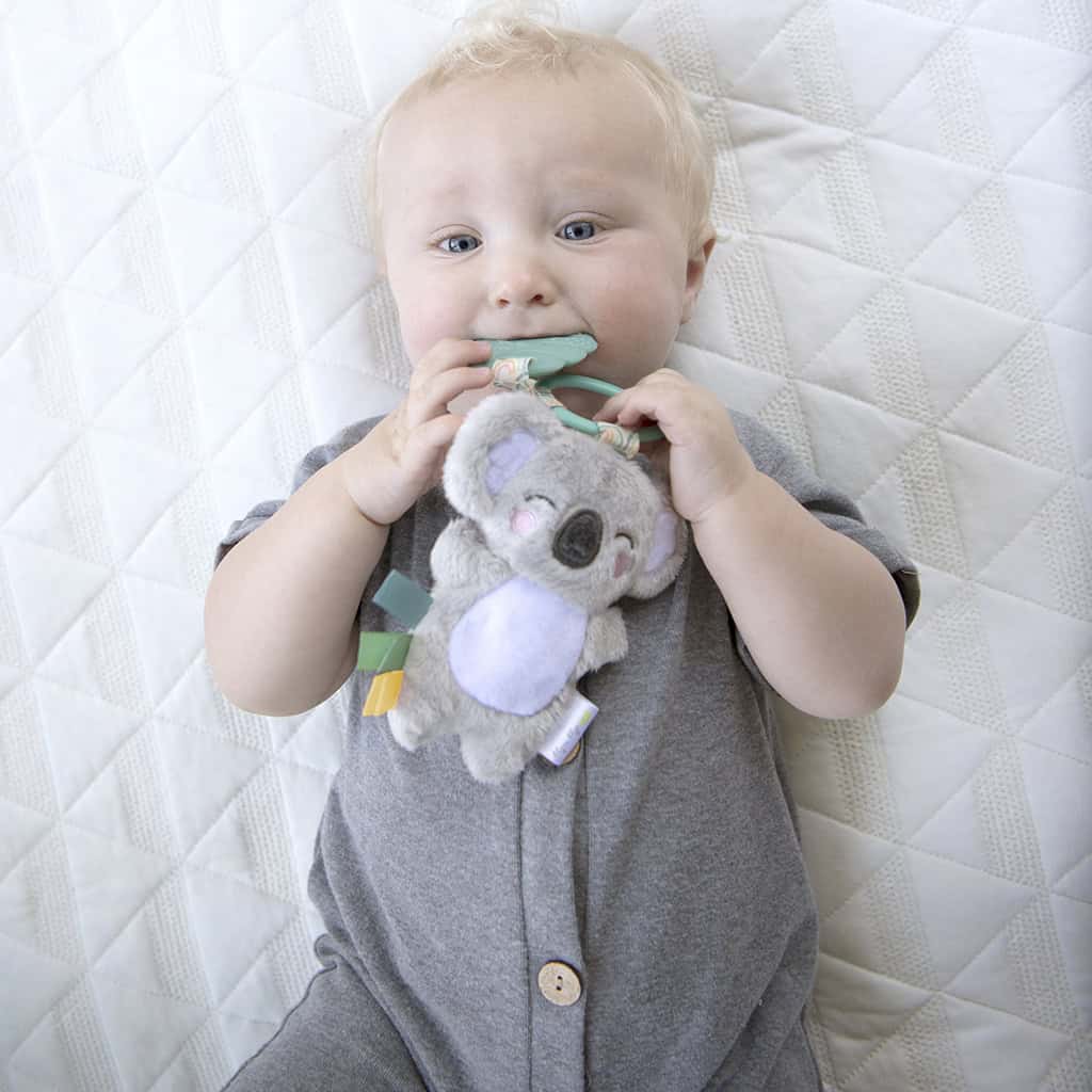 itzy-ritzy-tpl8443-itzy-pal-koala-1 A baby with blond hair, lying on a quilted blanket, happily chewing on an Itzy Ritzy - Itzy Pal - Plush Pal infant toy with silicone teether.