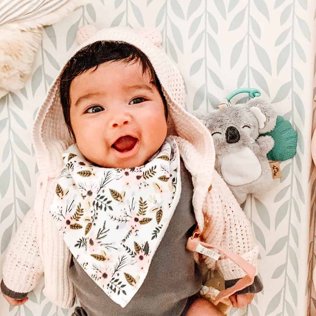 itzy-ritzy-tpl8443-itzy-pal-koala-5 A joyful baby with a floral bib and a bear-eared hood smiles widely, surrounded by soft toys and patterned wallpaper with the Itzy Ritzy - Itzy Pal - Plush Pal Infant Toy with Silicone Teether.