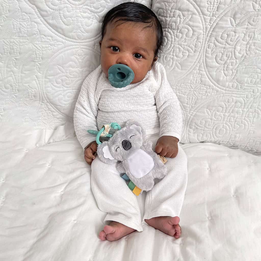 itzy-ritzy-tpl8443-itzy-pal-koala-8 A baby with a pacifier sits on a white quilt, holding an Itzy Ritzy - Itzy Pal - Plush Pal Infant Toy with Silicone Teether.