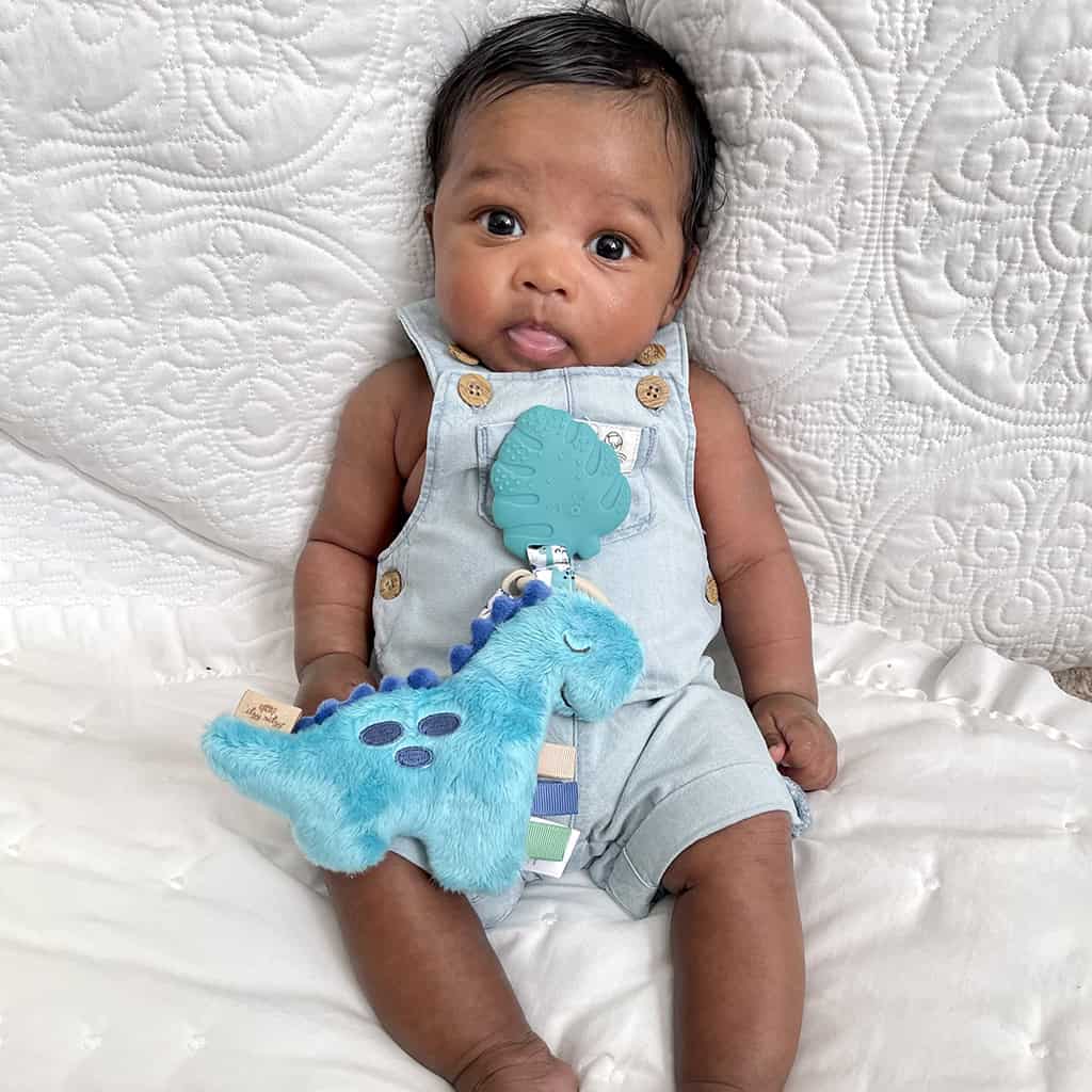 itzy-ritzy-tpl8449-itzy-pal-dino-2 Baby in a denim jumpsuit holding an Itzy Ritzy - Itzy Pal - Plush Pal Infant Toy with Silicone Teether, sitting on a white quilted background.