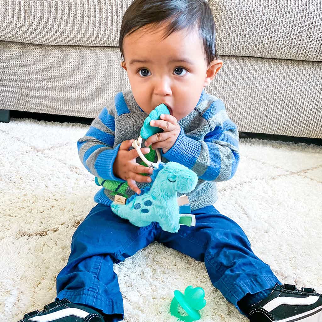 itzy-ritzy-tpl8449-itzy-pal-dino-6 Baby sitting on a white rug, chewing on an Itzy Ritzy - Itzy Pal - Plush Pal Infant Toy with Silicone Teether, wearing a striped sweater and jeans, with a pacifier nearby.