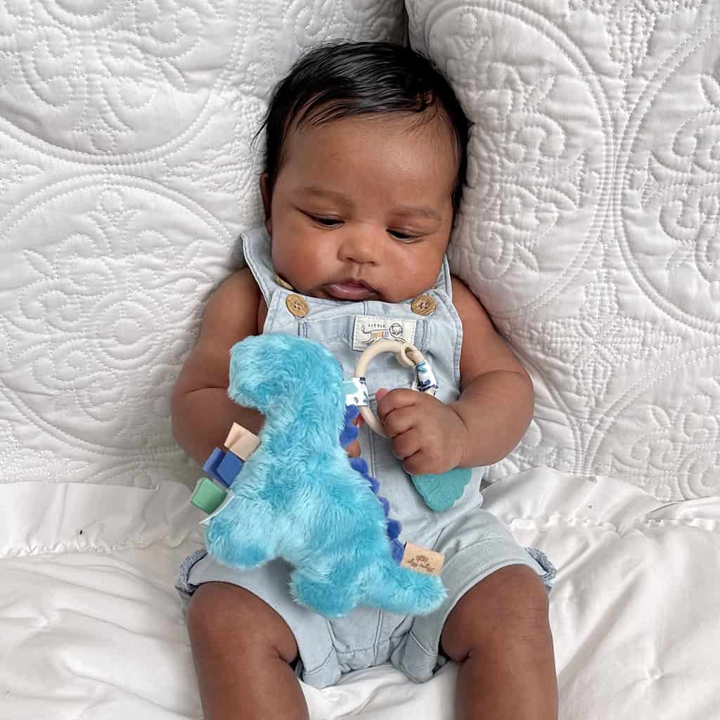 itzy-ritzy-tpl8449-itzy-pal-dino-7 A baby sitting on a textured blanket, holding an Itzy Ritzy - Itzy Pal - Plush Pal Infant Toy with Silicone Teether, dressed in a denim jumper with button details.